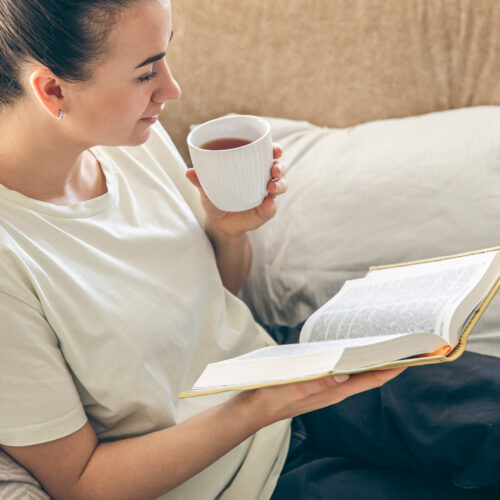 Young woman drinking hot tea, reading favorite paper book at home.